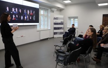 Prelegentka prowadzi prezentację w sali konferencyjnej, a uczestnicy siedzący na krzesłach oglądają slajd wyświetlany na dużym ekranie.