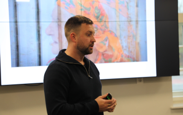 Mężczyzna z krótkim zarostem i ciemnymi włosami stoi z profilu, prowadząc prezentację. Ma na sobie ciemnogranatową bluzę. W tle widać duży ekran, na którym wyświetlany jest portret starszej osoby w kolorowej chustce na głowie.