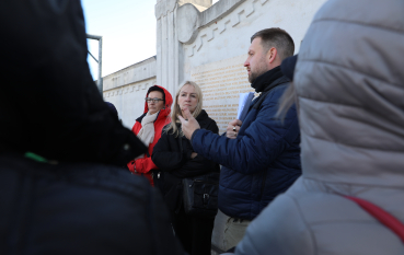 Mężczyzna z krótkim zarostem i ciemnymi włosami stoi z profilu i przemawia do grupy osób. Ma na sobie ciemnogranatową kurtkę, a w ręku trzyma plik kartek. ekran, na którym wyświetlany jest portret starszej osoby w kolorowej chustce na głowie.