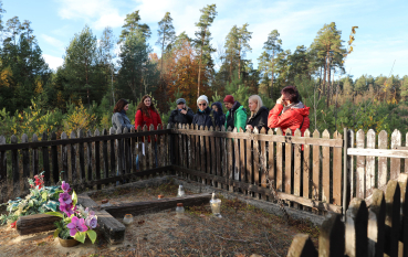 Grupa ośmiu osób stoi przy skromnym, otoczonym niskim drewnianym płotkiem grobie w lesie. Uczestnicy patrzą w stronę mogiły, na której leży drewniany krzyż, kwiaty oraz stoją znicze. W tle rozciąga się sosnowy las