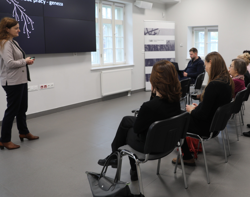 Na zdjęciu widać prelekcję w jasnej, nowoczesnej sali. Po lewej stronie stoi prowadząca przy dużym ekranie z napisem „obóz pracy – geneza”, a naprzeciwko niej siedzi grupa zasłuchanych osób.