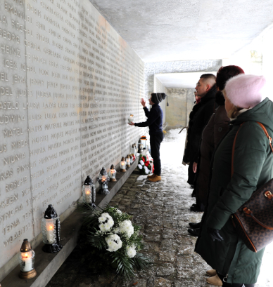 Perspektywiczne ujęcie betonowego muru pamięci, na którym wyryto imiona żydowskie. Przed ścianą stoi grupa osób w zimowych ubraniach, oddając hołd zmarłym. Chłopiec trzyma zapalony znicz i modli się. Na niskim podeście u podnóża muru gdzie leży również wiązanka białych kwiatów i palą się inne lampiony. 