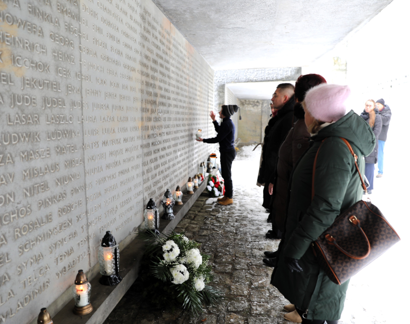 Perspektywiczne ujęcie betonowego muru pamięci, na którym wyryto imiona żydowskie. Przed ścianą stoi grupa osób w zimowych ubraniach, oddając hołd zmarłym. Chłopiec trzyma zapalony znicz i modli się. Na niskim podeście u podnóża muru gdzie leży również wiązanka białych kwiatów i palą się inne lampiony. 