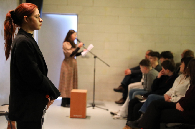 Młoda kobieta w czarnej marynarce stoi w skupieniu na pierwszym planie. W tle inna osoba przemawia do mikrofonu przed grupą siedzących ludzi w surowym, jasnym wnętrzu.