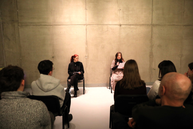 Dwie kobiety z mikrofonami siedzą przodem do publiczności na tle wysokiej, betonowej ściany, wykonując czytanie performatywne