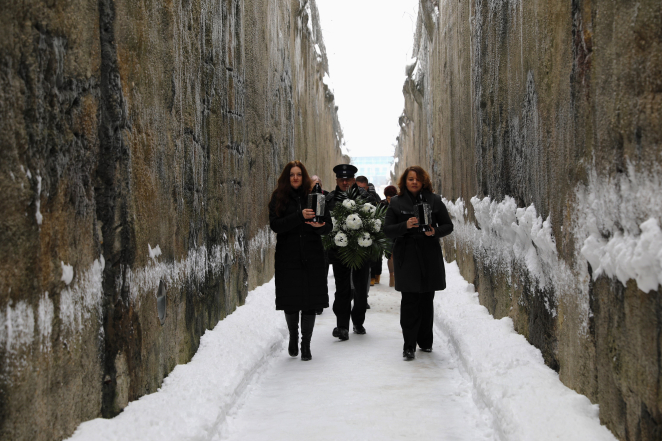 Wąski korytarz ośnieżonej ścieżki, ograniczony z obu stron wysokimi, ciemnymi ścianami o surowej, kamiennej fakturze. Na czele idą dwie kobiety w ciemnych, zimowych płaszczach, trzymając w dłoniach lampiony. Pomiędzy nimi idzie mężczyzna w mundurze, niosąc wieniec z białych kwiatów. Za nimi podąża grupa osób.