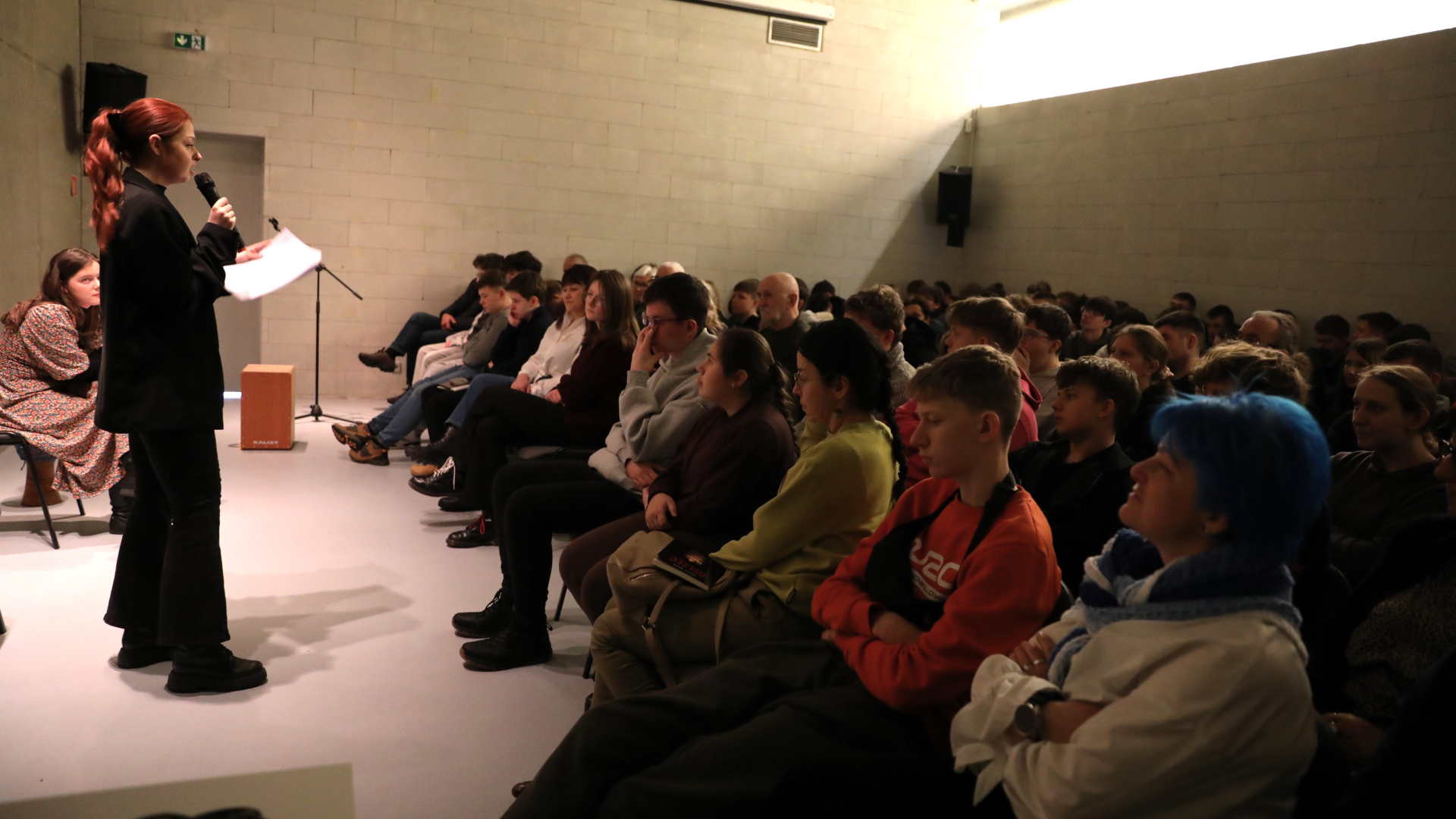 Ubrana na czarno kobieta trzymająca kartki i mikrofon stoi przed grupą siedzącej na krzesłach i przysłuchującej się grupie młodych osób.