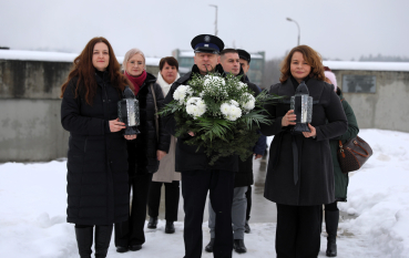 Grupa osób idących przez zaśnieżony teren. Na przedzie mężczyzna w mundurze niesie duży wieniec z białych kwiatów, a towarzyszące mu kobiety niosą zapalone znicze.