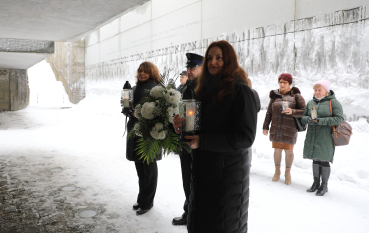 Delegacja ze zniczami i wieńcem idzie wzdłuż betonowej ściany z inskrypcjami. Wokół leży dużo śniegu.