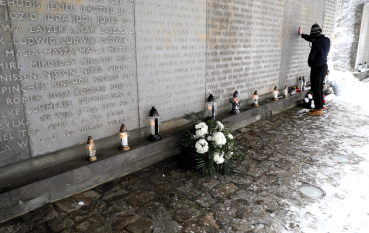 Ściana pamięci z wyrytymi imionami, pod którą złożono wieniec z białych kwiatów i liczne zapalone znicze. W tle widać młodego chłopca dotykającego dłonią muru.
