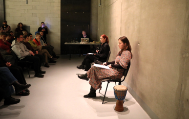 Dwie młode dziewczyny siedzą w minimalistycznej sali, naprzeciwko nich siedząca grupa osób