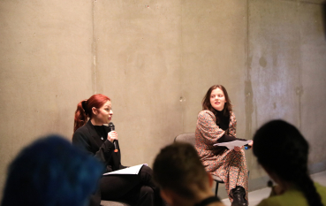 Zbliżenie na dwie kobiety siedzące na tle betonowej ściany; rudowłosa kobieta mówi do mikrofonu. W dolnej części kadru widoczne są nieostre sylwetki osób z publiczności