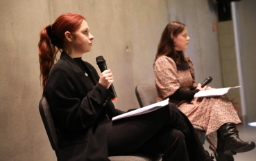 Profil młodej kobiety z rudymi włosami spiętymi w kucyk, która mówi do mikrofonu. W tle siedzi druga kobieta; obie znajdują się w surowym, betonowym pomieszczeniu.