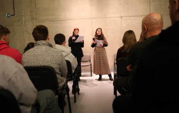 Widok zza pleców publiczności na dwie młode kobiety stojące przy betonowej ścianie. Obie trzymają mikrofony i kartki z tekstem.