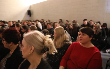 Zbliżenie na publiczność siedzącą w rzędach. Na pierwszym planie widać profile kobiet słuchających wystąpienia w stonowanym oświetleniu.