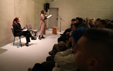 Szeroki kadr sali. Jedna kobieta siedzi, druga stoi i czyta z kartki do mikrofonu przed zgromadzoną publicznością. W tle widać minimalistyczne, industrialne wnętrze.