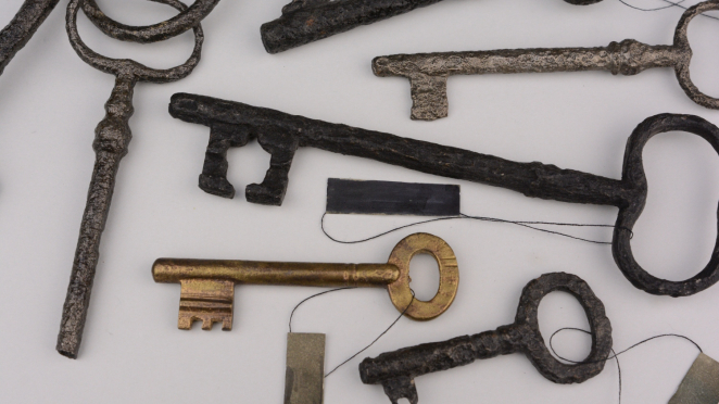 Kolekcja starych, w większości silnie skorodowanych kluczy na jasnym tle. Przedmioty mają etykiety inwentarzowe na nitkach, a wśród nich znajduje się jeden lepiej zachowany klucz w kolorze mosiądzu.