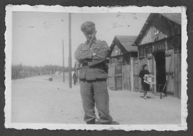 Na pierwszym planie stoi mężczyzna w niemieckim mundurze wojskowym - formacji SS, z rękami skrzyżowanymi na piersi i czapką nasuniętą na czoło. W tle widoczne są drewniane baraki. Po prawej stronie w głębi widać kobietę niosącą stołek. Po lewej stronie ciągnie się rząd drewnianych słupów, a podłoże jest piaszczyste i pozbawione roślinności.