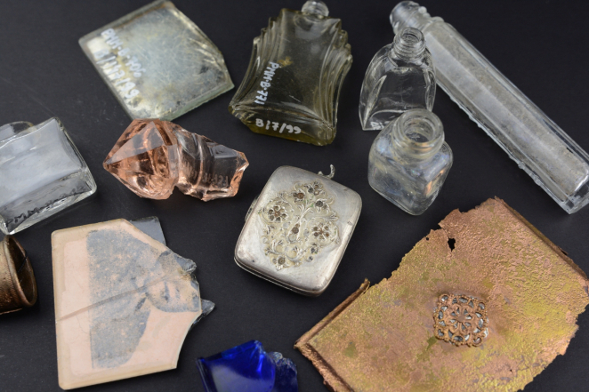 Drobne przedmioty na ciemnym tle: szklane flakoniki, fragmenty kolorowego szkła z fotografią dziecka, srebrna kasetka oraz skorodowana ozdobna blaszka z rozetą. 