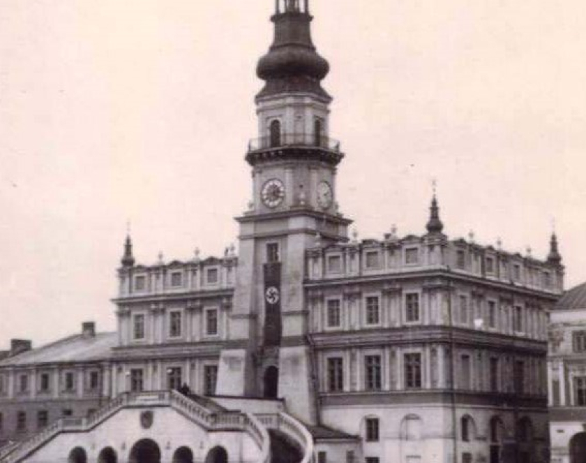 Czarno-białe zdjęcie przedstawia Ratusz w Zamościu w okresie okupacji niemieckiej podczas II wojny światowej. Na elewacji wieży, bezpośrednio nad wejściem, zawieszona jest pionowa flaga ze swastyką.