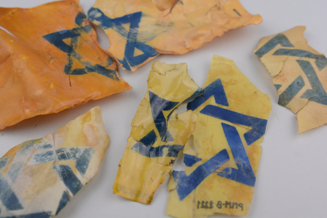 Sześć postrzępionych fragmentów żółtego materiału z nadrukowaną niebieską gwiazdą Dawida, ułożonych na jasnym tle. Na jednym z kawałków widoczny jest odręczny numer inwentarzowy.