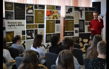 Po prawej stronie stoi mężczyzna przemawiający do grupy osób siedzących w rzędach tyłem do obiektywu. W tle znajdują się cztery wysokie banery wystawowe z nagłówkiem „Niemiecki obóz zagłady w Bełżcu. Historia i relikty, prezentujące zdjęcia i opisy przedmiotów oraz dokumentów historycznych.