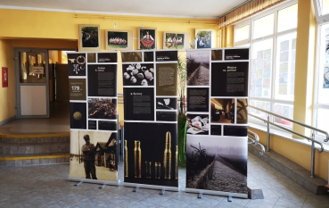 Trzy wysokie banery wystawowe ustawione obok siebie w jasnym holu o żółtych ścianach. W tle widoczne są schody, przeszklone drzwi oraz mniejsze, kolorowe zdjęcia zawieszone na ścianie.