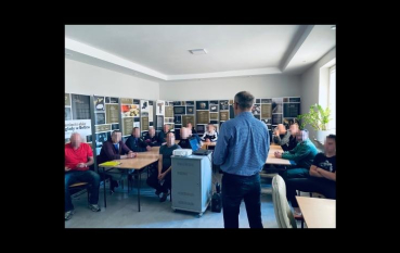 Zdjęcie dokumentuje spotkanie edukacyjne odbywające się w jasnej sali. Prowadzący, stoi tyłem do obiektywu i przodem do grupy kilkunastu mężczyzn siedzących przy stołach. Twarze uczestników zostały zamazane. Przy ścianach znajdują się planszami wystawy.