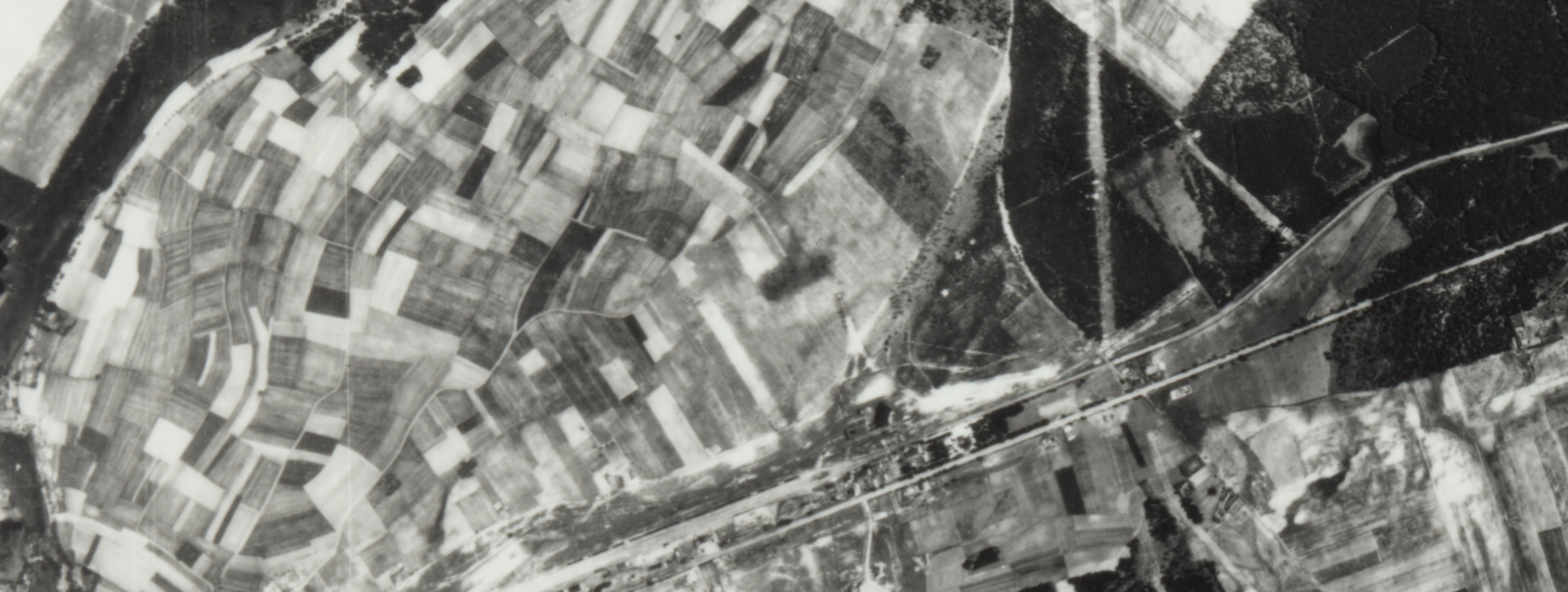 Czarno-białe zdjęcie lotnicze przedstawiające rozległy obszar wiejski i leśny. Widok z góry ukazuje mozaikę pól uprawnych, kompleksy leśne oraz przebiegające przez środek linie komunikacyjne.