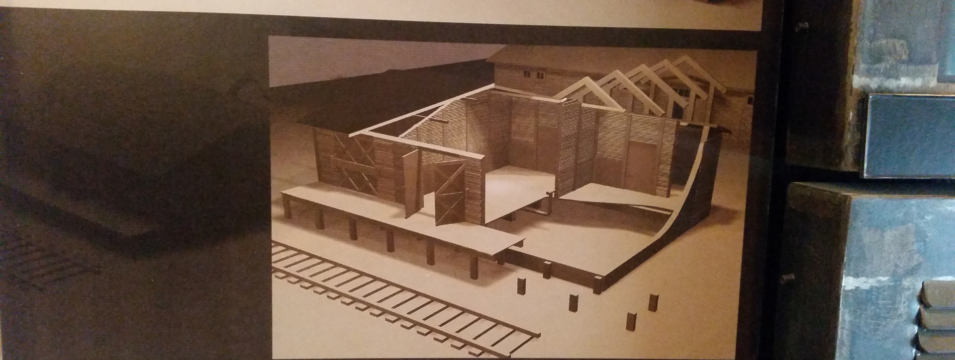 Grafika przedstawia drewnianą konstrukcję o charakterze barakowym, pokazaną bez dachu i części ścian zewnętrznych, co pozwala zajrzeć do wnętrza. Widoczne są wewnętrzne przegrody, belki konstrukcyjne oraz podwyższony podest wsparty na słupach.