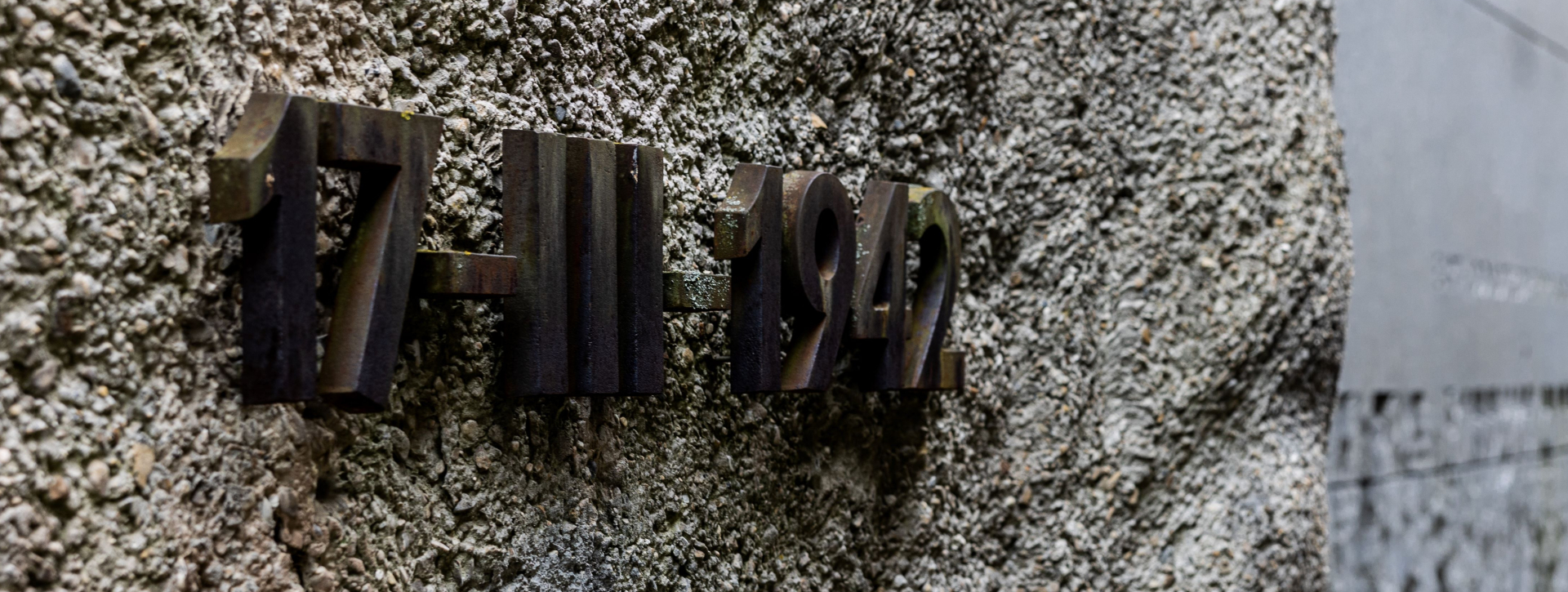 Zdjęcie przedstawia zbliżenie na metalową datę 17-3-1942 umieszczoną na chropowatej, betonowej ścianie pomnika. W rozmazanym tle widać ścianę oraz złożone na ziemi kwiaty.