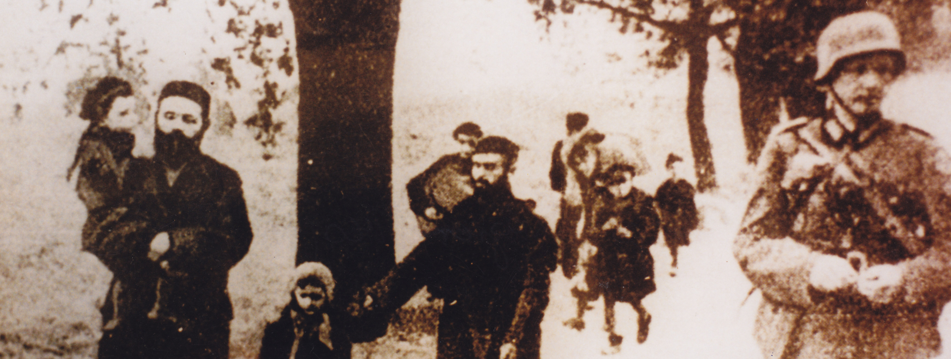 Archiwalne zdjęcie przedstawiające kolumnę cywilów, w tym mężczyźni w długich płaszczach z dziećmi, idący drogą wzdłuż drzew. Po prawej stronie, na pierwszym planie, maszeruje uzbrojony żołnierz w hełmie.