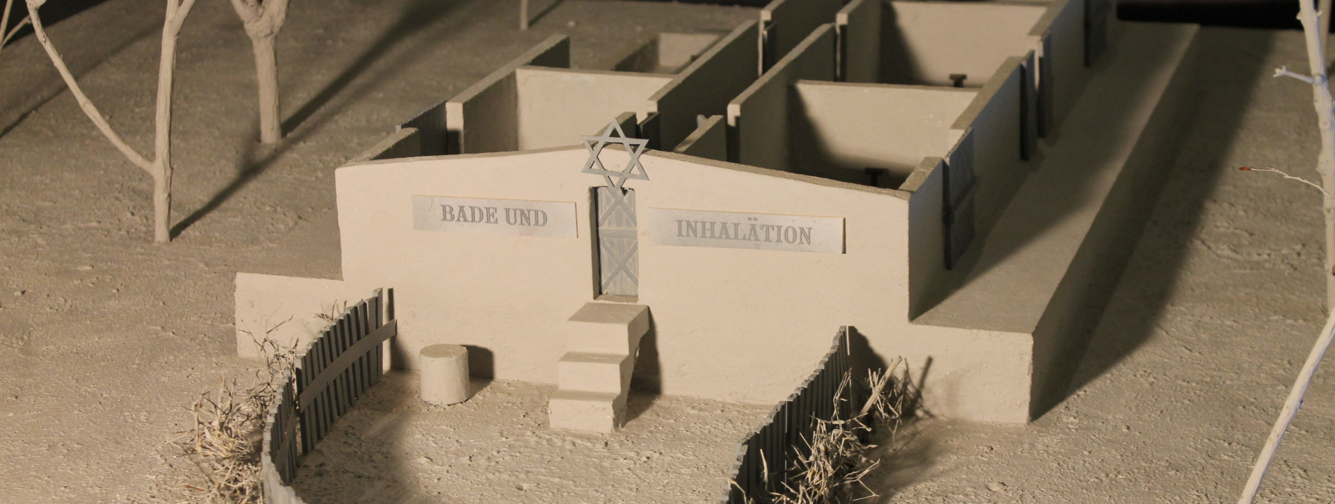 Zdjęcie przedstawia makietę budynku komór gazowych. Model jest wykonany w jasnoszarym kolorze i nie posiada dachu, co ukazuje podział wnętrza na kilka mniejszych, prostokątnych pomieszczeń. Na frontowej ścianie znajdują się centralne drzwi, do których prowadzą schodki. Nad wejściem umieszczono symbol Gwiazdy Dawida, a po jego bokach widnieją tablice z niemieckimi napisami: „BADE UND” (po lewej) oraz „INHALATION” (po prawej). Przed budynkiem widoczne jest niskie, drewniane ogrodzenie. W tle makiety widać suche gałęzie drzew.