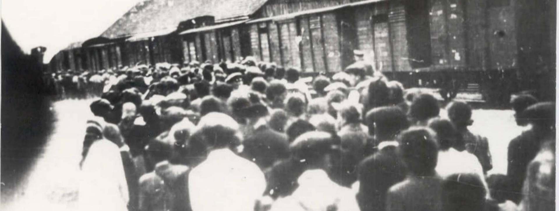 Historyczne zdjęcie przedstawiające tłum ludzi zgromadzonych na peronie obok pociągu towarowego z drewnianymi wagonami. Scena dokumentuje deportację ludności w czasie wojny, w drodze do obozu zagłady.