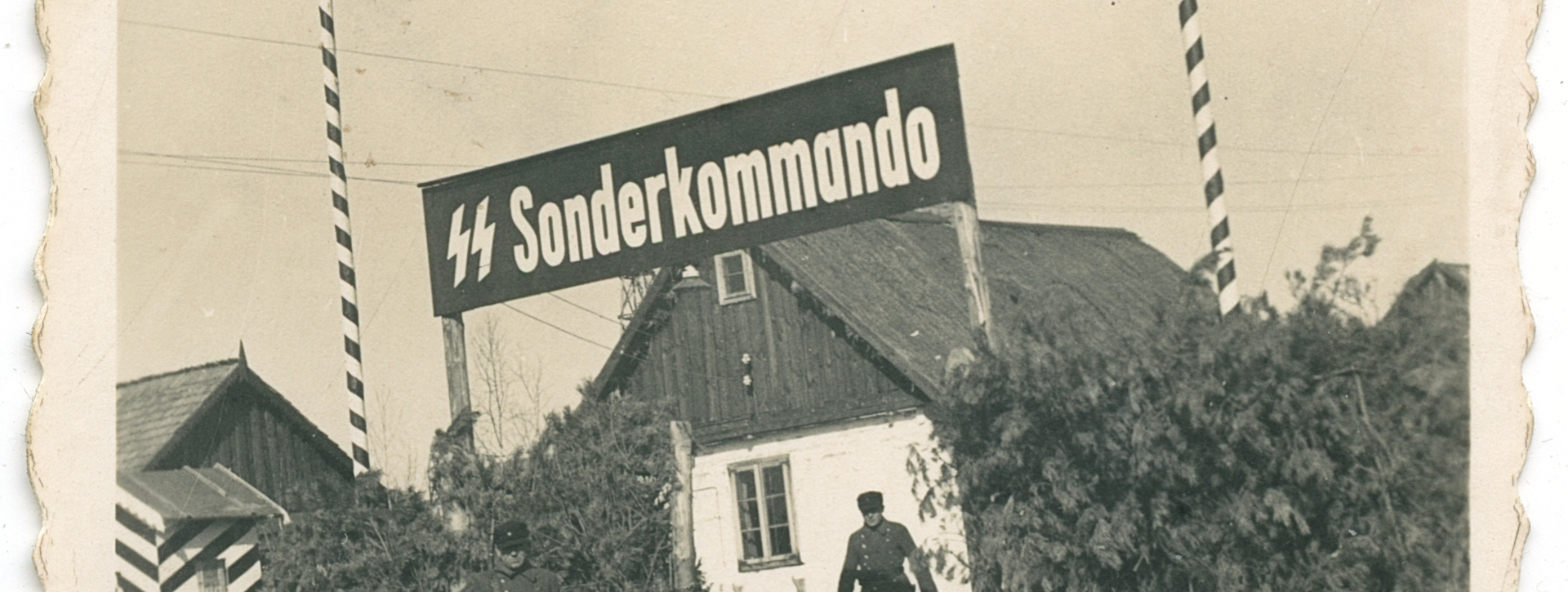 Historyczne zdjęcie przedstawiające bramę wjazdową do obozu zagłady z widocznym napisem „SS Sonderkommando”. Przy wejściu powiewają flagi III Rzeszy i SS, a dostępu bronią strażnicy w mundurach. Całość otoczona jest wysokim żywopłotem maskującym.