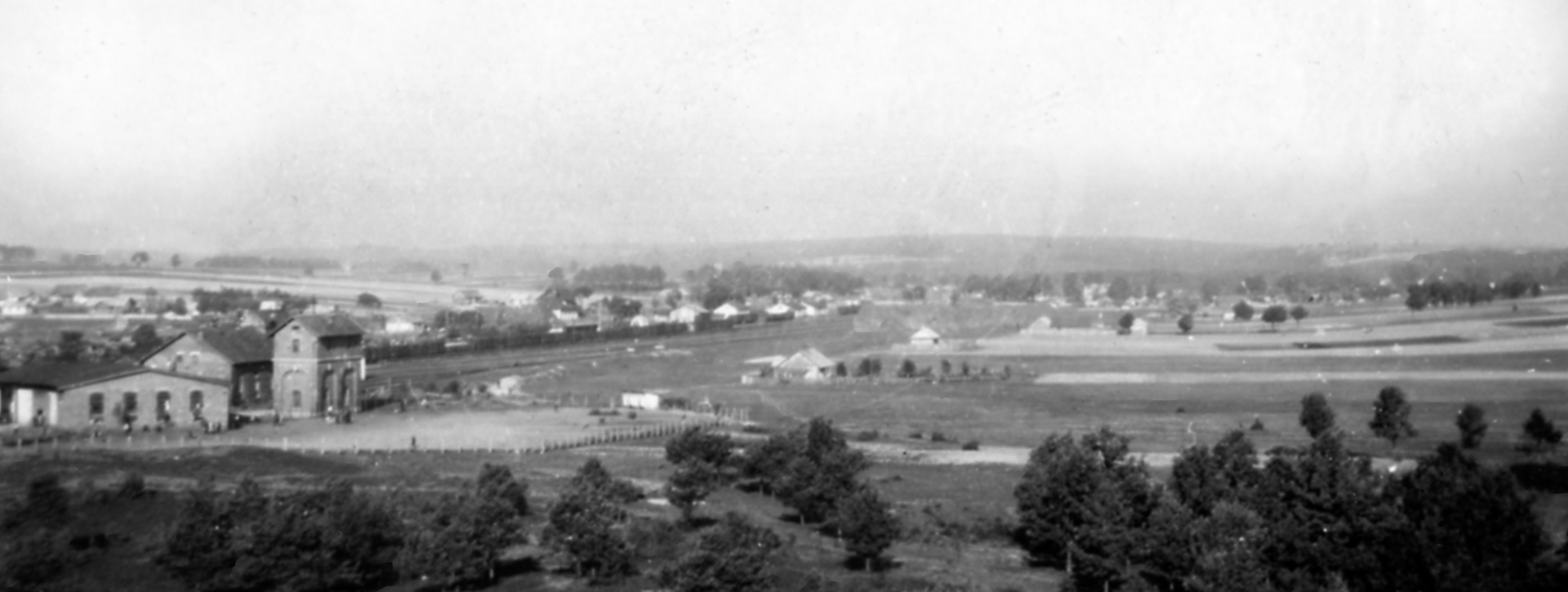 Czarno-białe, archiwalne zdjęcie panoramiczne przedstawiające rozległy krajobraz z niską zabudową w oddali. Na pierwszym planie po lewej stronie widoczne są murowane budynki, a resztę terenu zajmują pola, kępy drzew i otwarta przestrzeń pod jasnym niebem.