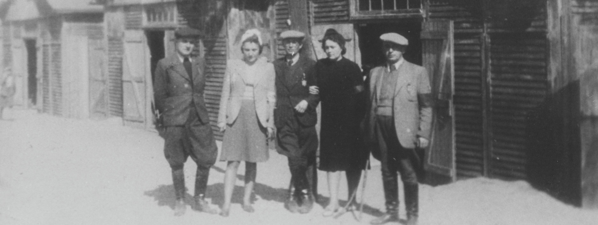 Czarno-biała fotografia przedstawiająca grupę pięciu osób — trzech mężczyzn i dwie kobiety — stojących na piaszczystym placu przed rzędem drewnianych baraków. Mężczyźni noszą marynarki, płaskie czapki (kaszkiety) i wysokie buty, a kobiety mają na sobie spódnice, marynarki i nakrycia głowy. U czterech osób na lewych ramionach widoczne są wyraźne opaski. W tle znajdują się identyczne drewniane budynki o konstrukcji z pionowych desek i dwuspadowych dachach. 