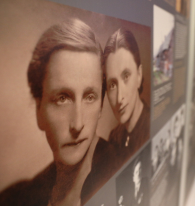 Czarno-białe, archiwalne zdjęcie dwóch kobiet o poważnych twarzach i gładko zaczesanych włosach, stanowiące główny element muzealnej wystawy. Portret jest umieszczony na ciemnej tablicy obok innych fotografii i tekstów historycznych, a całe ujęcie wykonano pod kątem, co nadaje kompozycji głębi.