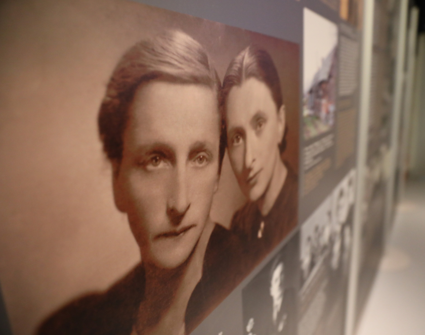 Czarno-białe, archiwalne zdjęcie dwóch kobiet o poważnych twarzach i gładko zaczesanych włosach, stanowiące główny element muzealnej wystawy. Portret jest umieszczony na ciemnej tablicy obok innych fotografii i tekstów historycznych, a całe ujęcie wykonano pod kątem, co nadaje kompozycji głębi.