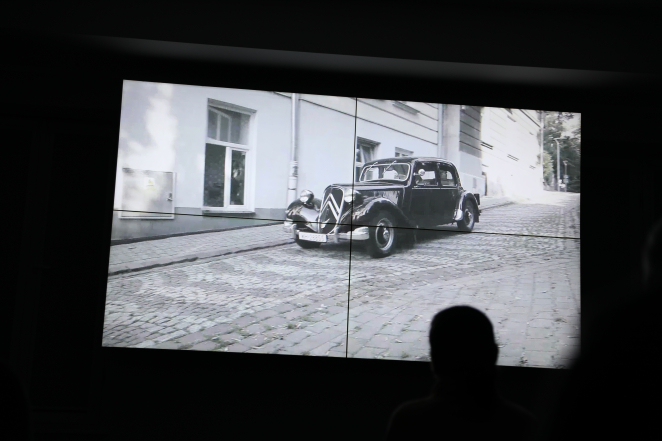 Czarno-biała projekcja na ekranie, przedstawiająca zabytkowy samochód marki Citroën zaparkowany na pochyłej, brukowanej ulicy. Na pierwszym planie, w przyciemnionej sali wystawowej, widoczna jest z tyłu sylwetka osoby obserwującej nagranie.