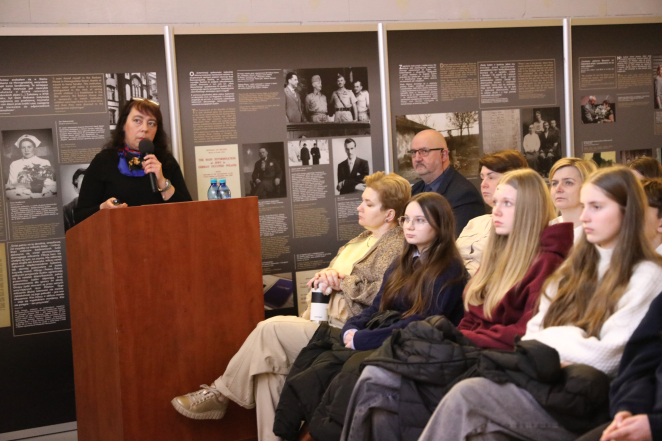 Kobieta przemawiająca do mikrofonu przy drewnianym pulpicie. Obok siedzi grupa zasłuchanych osób, a w tle znajdują się duże tablice edukacyjne z archiwalnymi fotografiami i tekstami historycznymi.