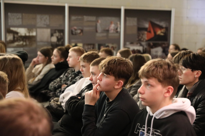 Publiczność siedząca w rzędach i uważnie słuchająca prelekcji podczas wydarzenia. W tle widoczne są panele ekspozycyjne z tekstami i zdjęciami - historie Sprawiedliwych.