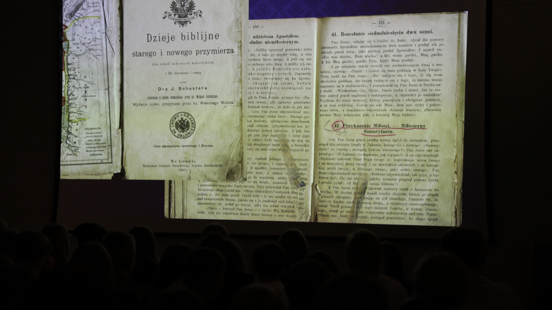 Projekcja skanów stron starej książki zatytułowanej „Dzieje biblijne starego i nowego przymierza” na dużym ekranie. W dolnej części zdjęcia widać zarysy głów publiczności siedzącej w zaciemnionej sali.