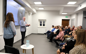 Mężczyzna w jasnoniebieskiej marynarce gestykuluje podczas przemówienia do grupy osób siedzących w rzędach.