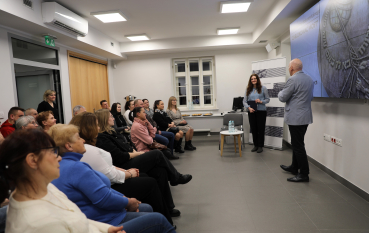 Widok na salę konferencyjną z profilu; prelegenci stoją przy ekranie, a publiczność uważnie słucha wprowadzenia.