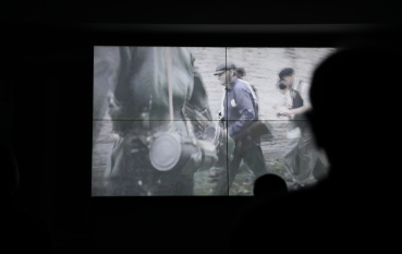 Zbliżenie na ekran, na którym wyświetlany jest czarno-biały fragment filmu dokumentalnego przedstawiający postacie w historycznych strojach.