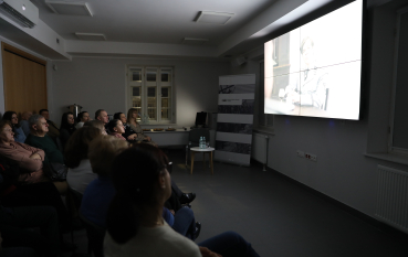 Publiczność w przyciemnionej sali ogląda film dokumentalny wyświetlany na dużym ekranie ściennym.