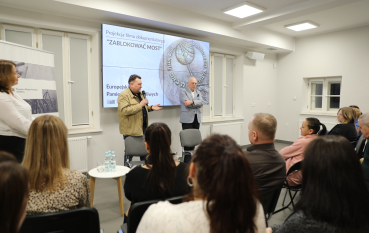 Mężczyzna w beżowej kurtce mówi do mikrofonu, stojąc obok drugiego prelegenta przed ekranem tytułowym filmu.