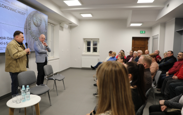 Dwaj mężczyźni prowadzą dyskusję przed siedzącą grupą osób; jeden z nich trzyma mikrofon, drugi słucha w zamyśleniu.