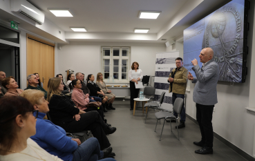 Mężczyzna w błękitnej marynarce energicznie przemawia i gestykuluje przed siedzącą publicznością w jasnej sali konferencyjnej.